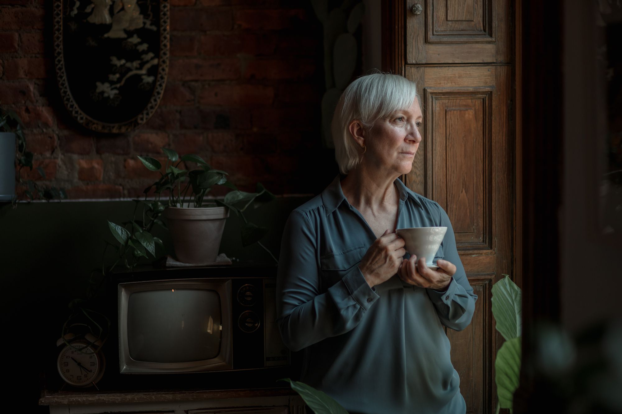 A person holding a cup of coffee at a window, looking out in soft morning light.